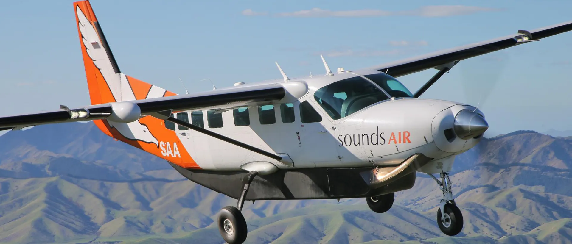 A white and orange Sounds Air turboprop plane flying over green, rolling hills with its landing gear down.