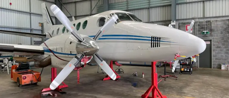 A white twin-engine turboprop aircraft with blue stripes, elevated on red maintenance jacks inside a large corrugated metal hangar.